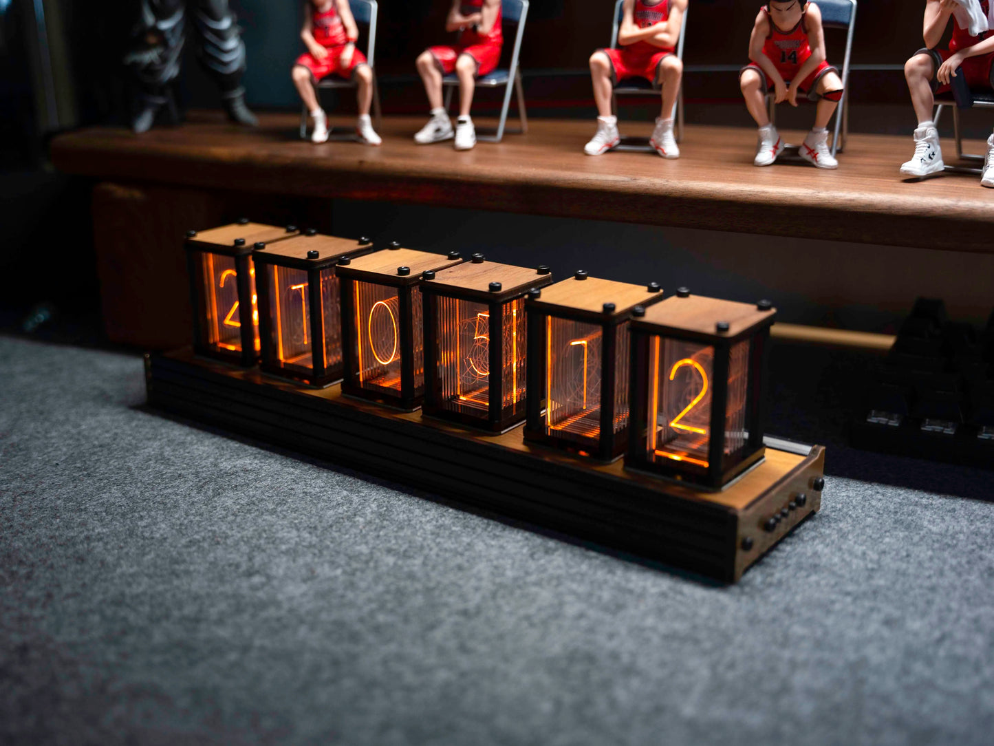 Digital clock with glowing numbers on a dark surface, blurred background of people sitting.
